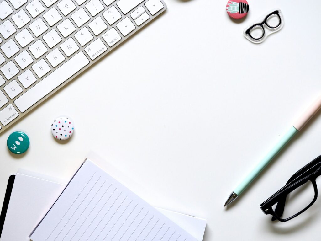 Carnet, clavier blanc, stylo menthe et lunettes disposés sur un bureau blanc.