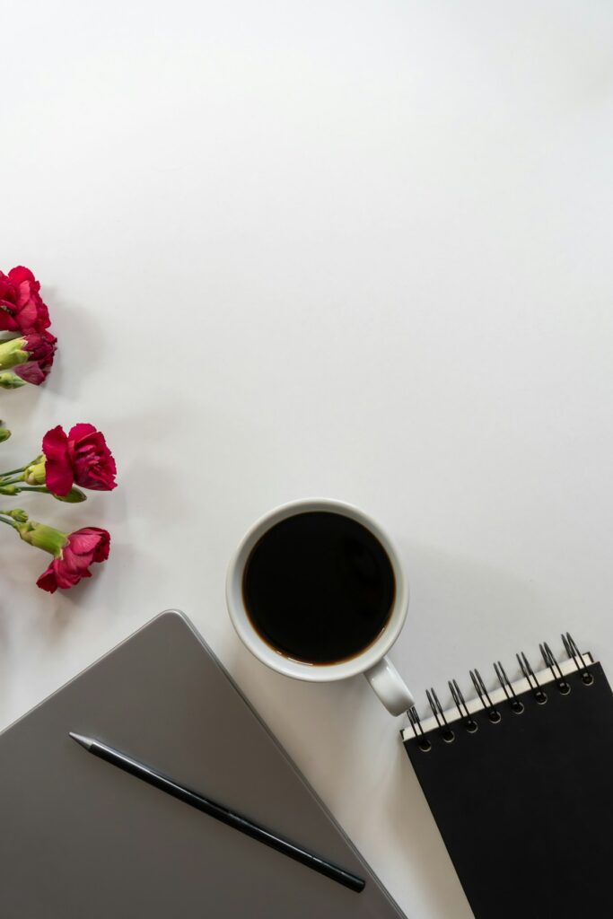 Ordinateur, tasse de café, carnet et petites fleurs rouges sur une surface claire.