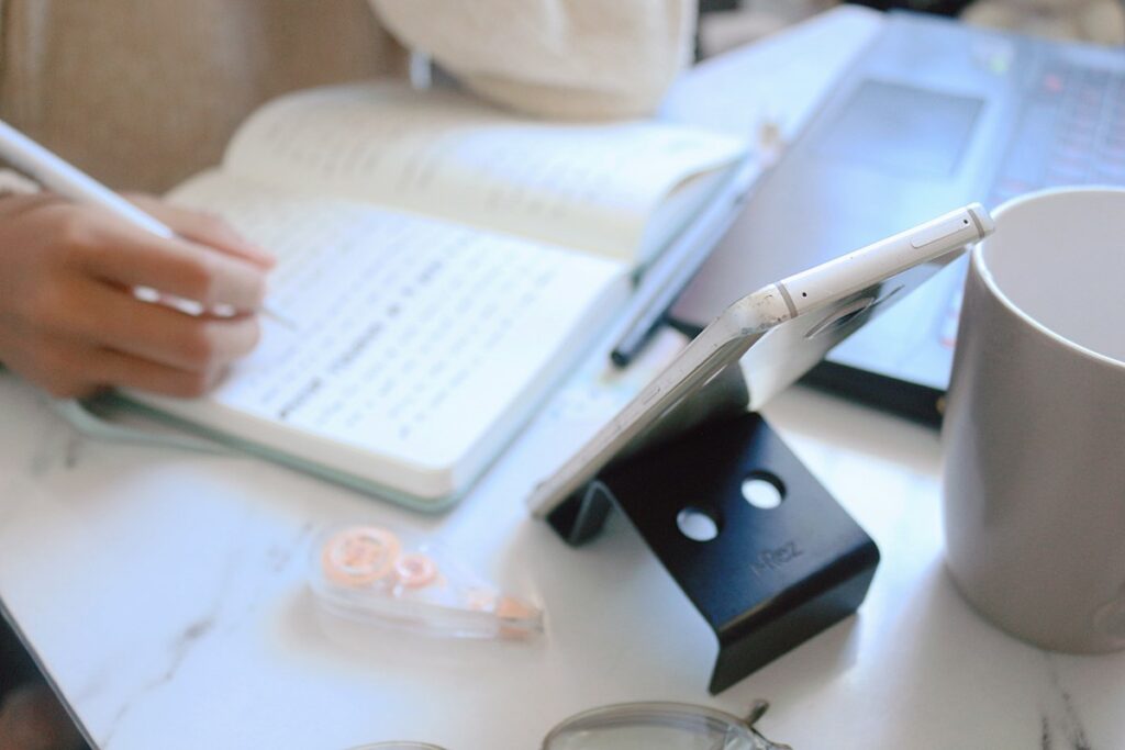 Main écrivant dans un carnet, téléphone et tasse sur un bureau blanc.