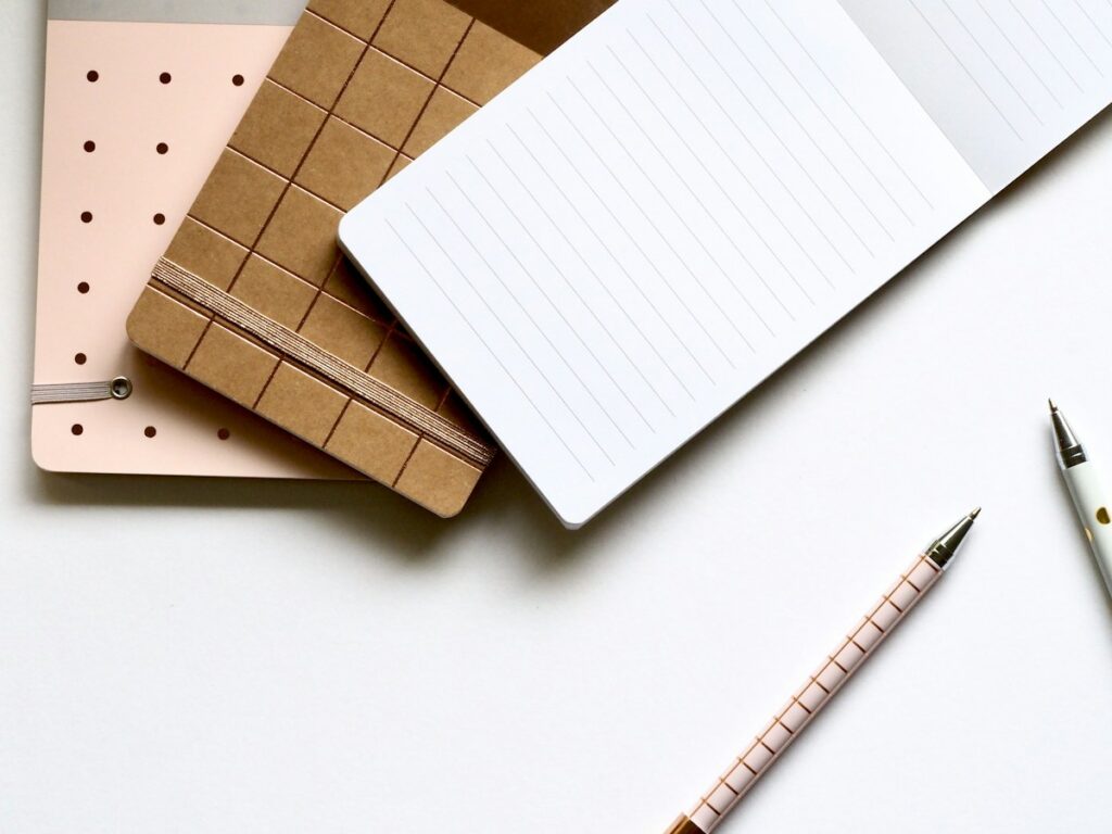 Carnets blancs et kraft, règle et stylo rose pâle disposés sur une surface blanche.