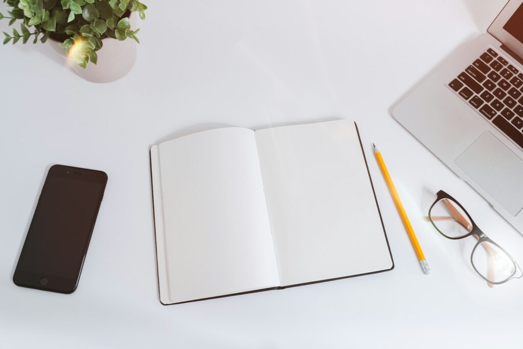 Carnet ouvert, ordinateur et téléphone disposés sur un bureau blanc.